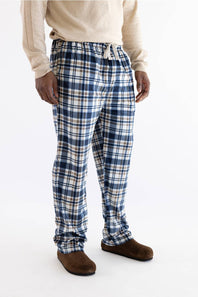 A person wearing blue, white, and beige plaid lounge pants with a drawstring waist, paired with a cream sweater and brown slippers, photographed against a plain white studio background.