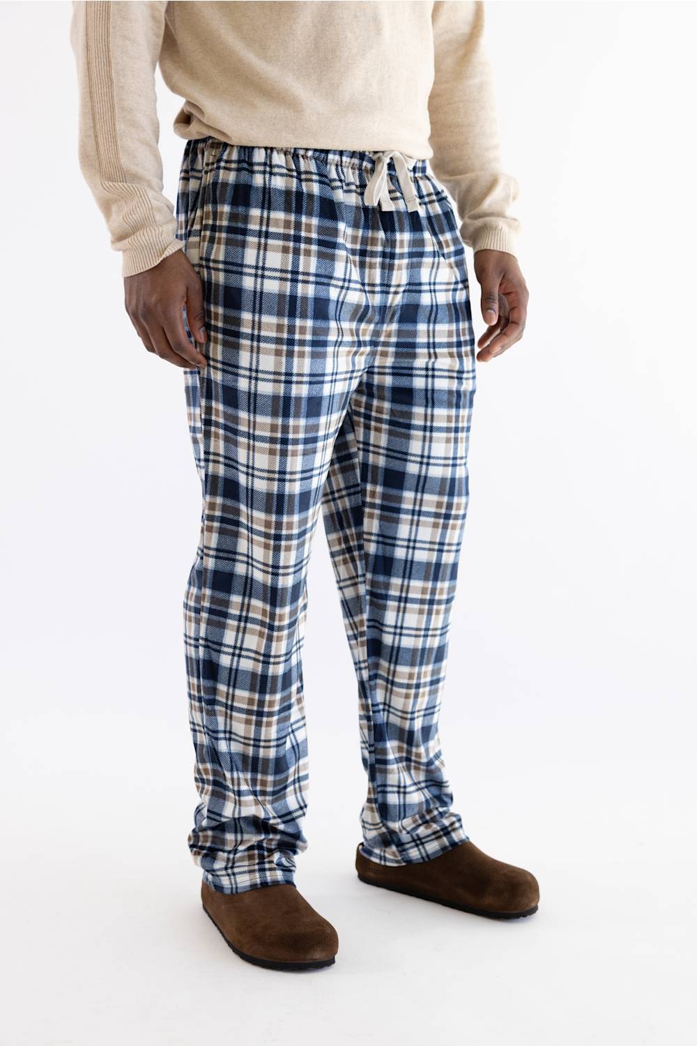 A person wearing blue, white, and beige plaid lounge pants with a drawstring waist, paired with a cream sweater and brown slippers, photographed against a plain white studio background.