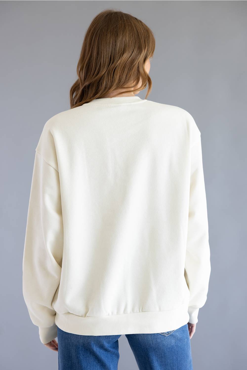 A person shown from the back wearing an oversized cream sweatshirt and blue jeans against a plain gray studio background.