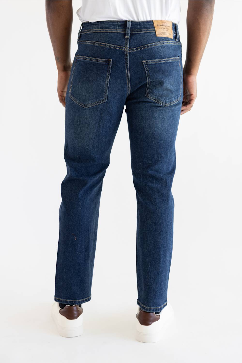 Rear view of a person wearing dark blue straight-leg jeans with a white t-shirt and white sneakers, photographed against a clean white studio background.