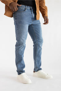 A person wearing light blue slim-fit jeans, a black top, a brown jacket, and white sneakers, shown from the chest down against a plain light background.