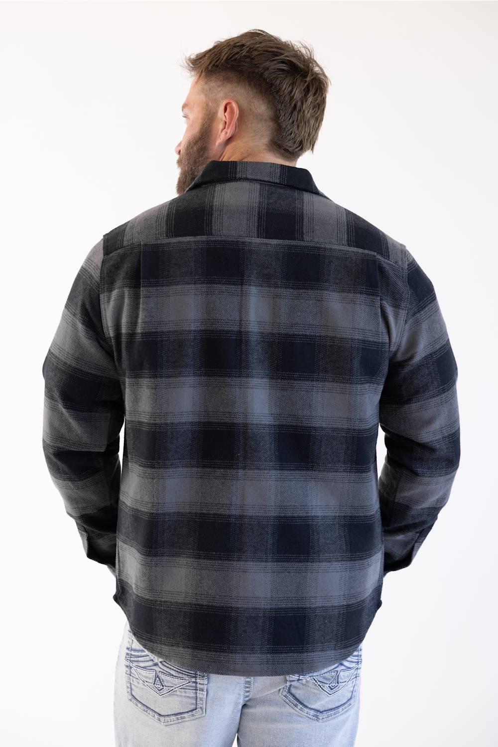 The back view of a man modeling the Weatherproof Vintage Lumberjack Plaid Shirt Jacket in "Phantom Night" (charcoal and dark navy plaid) worn over light-wash jeans, seen from behind as he looks slightly to his left, against a white studio background.
