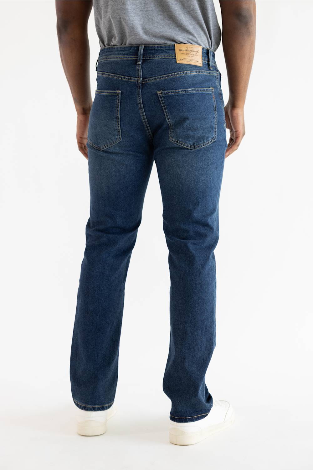 Rear view of a person wearing dark blue straight-leg jeans with a gray t-shirt and white sneakers, photographed against a clean white studio background.