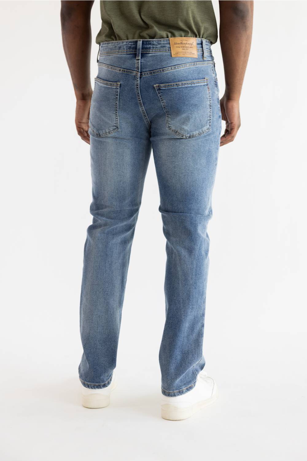 Rear view of a person wearing light blue denim jeans with a green shirt and white sneakers against a plain white studio background.