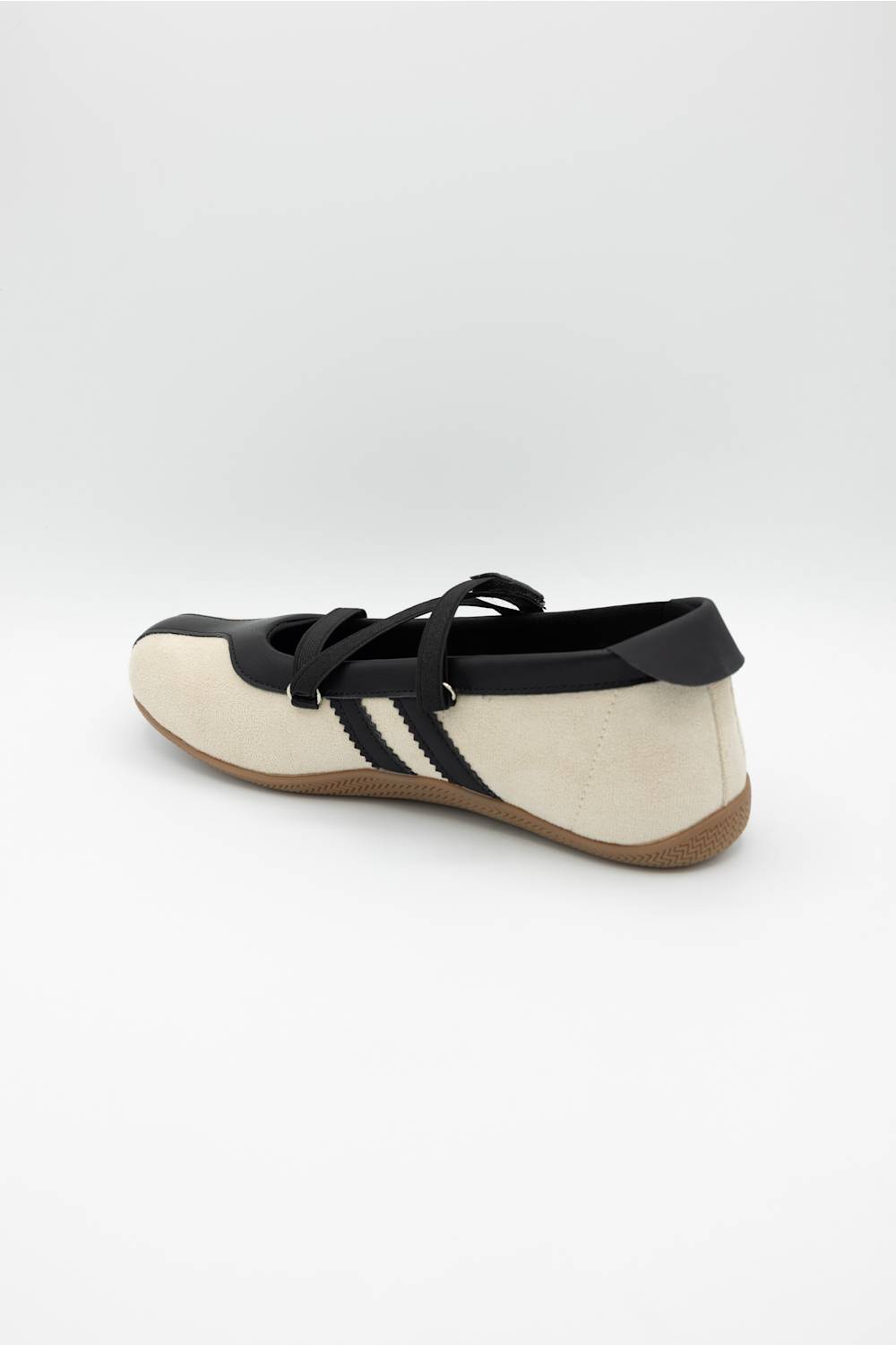 A single low-top sneaker shown from a side-rear angle on a light gray studio background. The shoe has a cream suede-like upper with black overlays, black laces, black side stripes, and a brown gum sole.