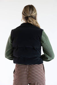 A woman viewed from behind wearing a black puffer vest over an olive green long-sleeve shirt and brown striped pants, against a plain white background.