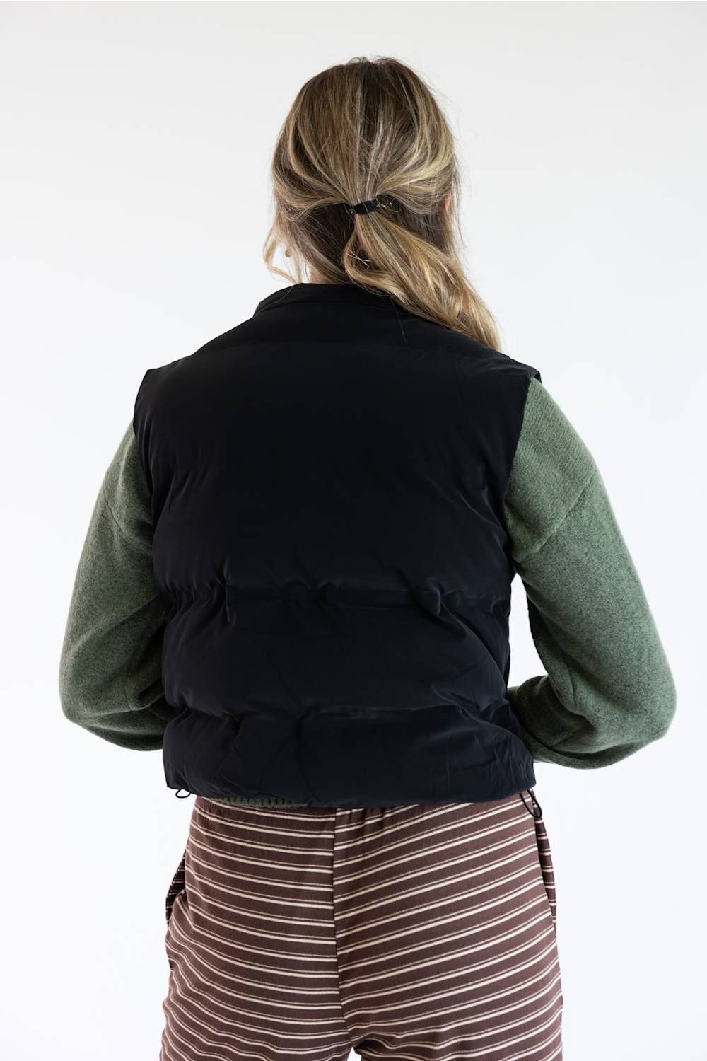 A woman viewed from behind wearing a black puffer vest over an olive green long-sleeve shirt and brown striped pants, against a plain white background.