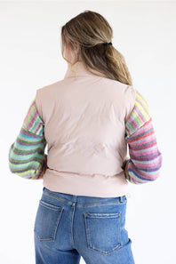 A woman shown from the back wearing a light pink puffer vest over a long-sleeve knit top with multicolored striped sleeves, paired with blue jeans, against a plain white background.