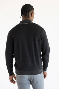 A man viewed from behind wearing a dark charcoal gray knit quarter-zip sweater with a contrasting light gray collar trim, paired with light gray trousers, against a plain white studio background.