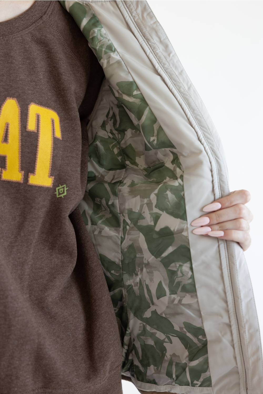 A close-up of a person wearing a brown sweatshirt with yellow lettering and holding open a light beige zippered jacket that reveals a green camouflage-patterned lining.