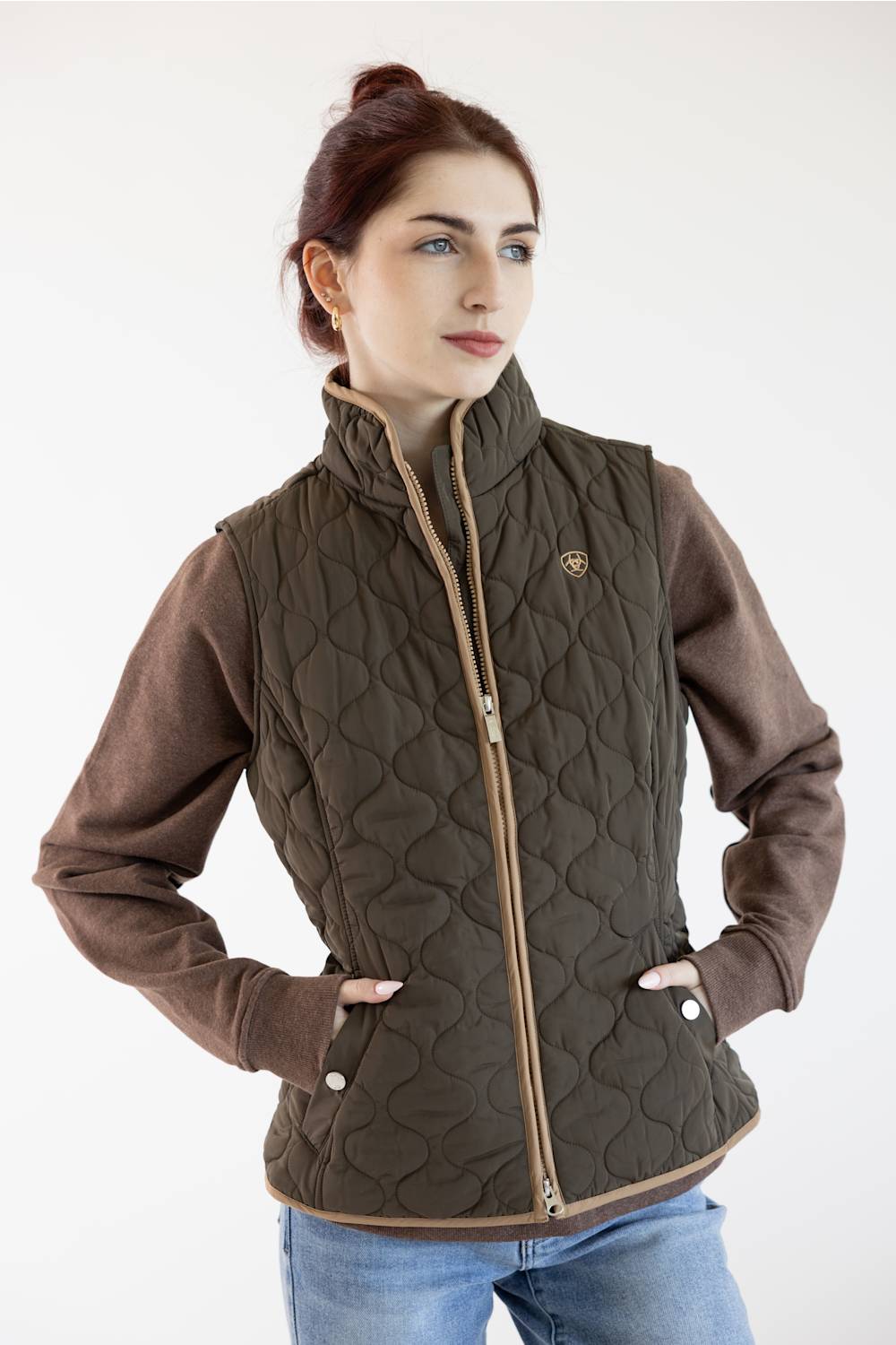 A woman wearing a dark brown quilted vest with brown sleeves and light brown trim, paired with blue jeans, against a plain white background.