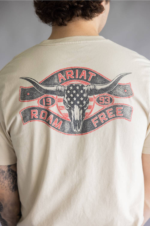 A person shown from the back wearing a cream-colored t-shirt with a large distressed graphic on the back. The graphic features a longhorn skull with an American flag pattern, flanked by banners reading 'ARIAT' and 'ROAM FREE' with '1993' in the center.
