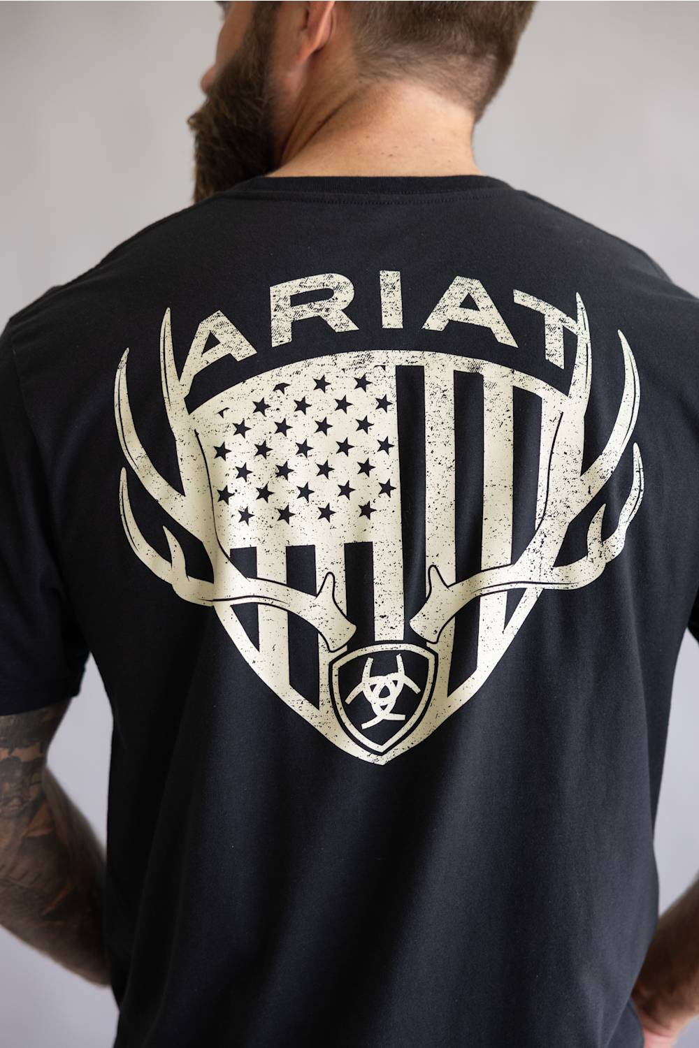 Back view of a man wearing a black t-shirt featuring a large distressed graphic of an American flag inside a shield with deer antlers and the Ariat brand name.