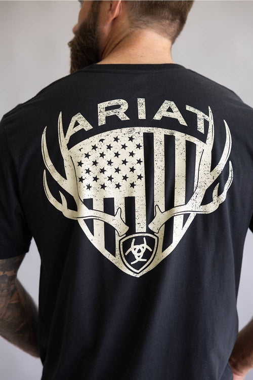 Back view of a man wearing a black t-shirt featuring a large distressed graphic of an American flag inside a shield with deer antlers and the Ariat brand name.