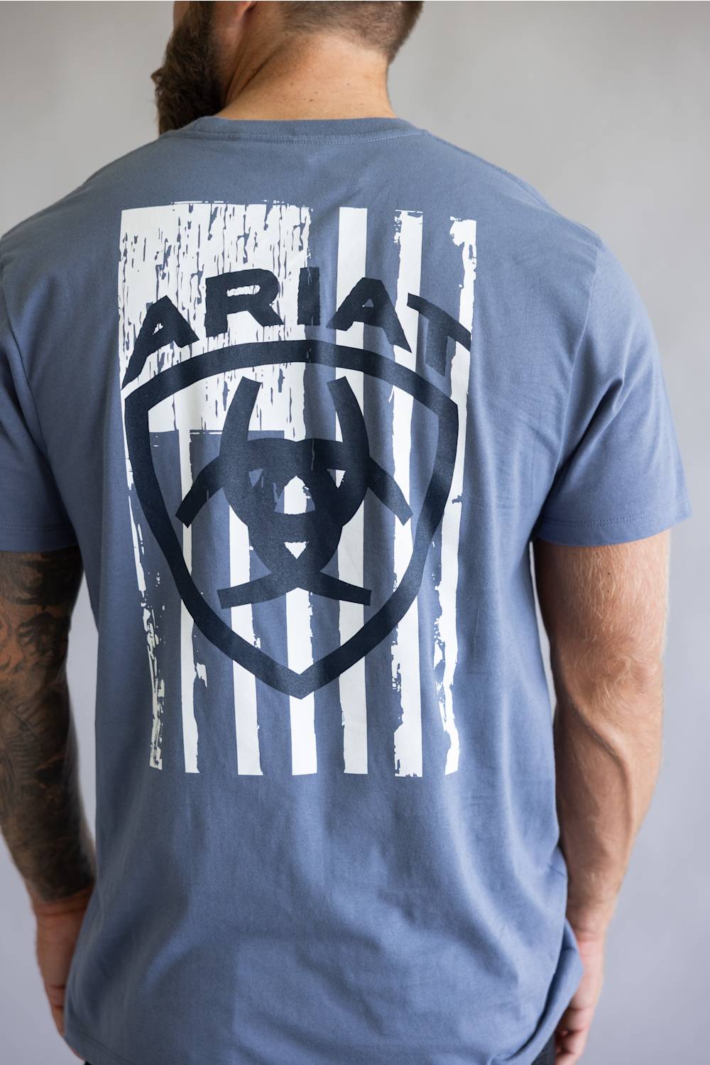 Back of a slate blue t-shirt featuring a distressed white American flag logo with the Ariat brand name.