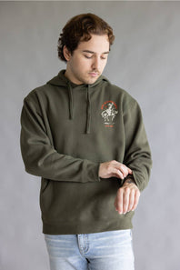 A man wearing an olive green pullover hoodie with a graphic print on the left chest, paired with light blue jeans, standing against a neutral gray background.