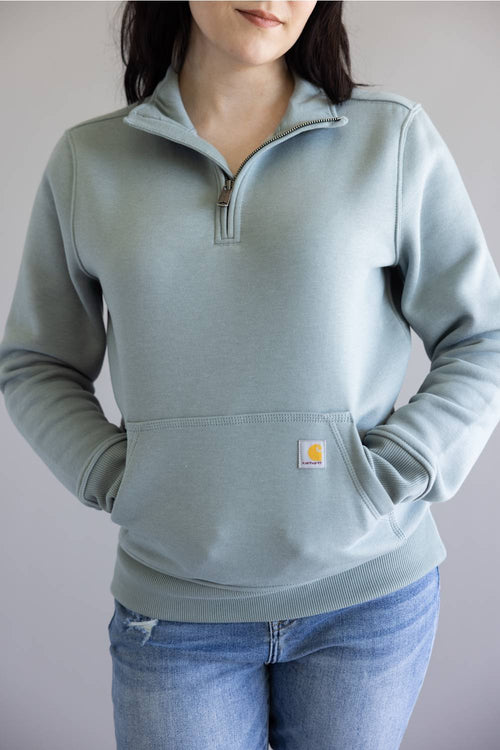 A woman wearing a light sage green Carhartt quarter-zip pullover sweatshirt with a kangaroo pocket and a small yellow Carhartt logo patch, paired with blue jeans.