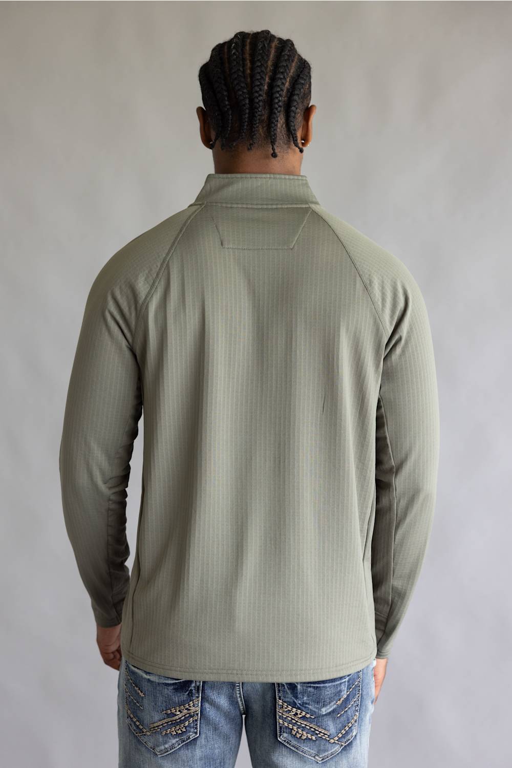 A man viewed from behind wearing a sage green ribbed long-sleeve quarter-zip pullover and blue jeans with decorative stitching on the back pockets, set against a plain gray studio background.