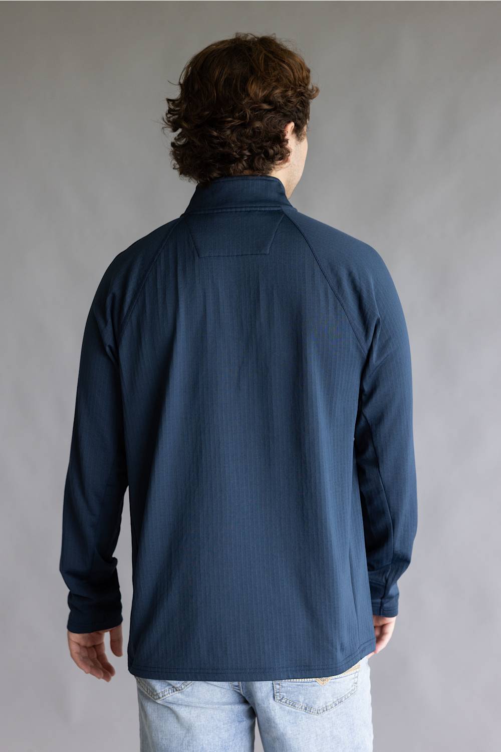 A man viewed from behind wearing a dark navy blue long-sleeve quarter-zip pullover shirt and light-wash jeans, standing against a neutral gray studio background.