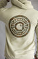 A person wearing a light beige hoodie from the back, featuring a large circular Carhartt logo printed in camouflage and white on the back.
