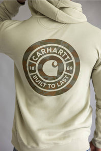 A person wearing a light beige hoodie from the back, featuring a large circular Carhartt logo printed in camouflage and white on the back.