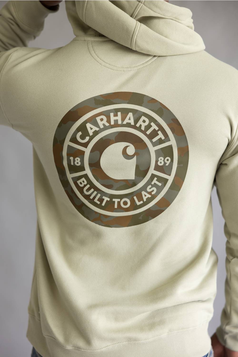 A person wearing a light beige hoodie from the back, featuring a large circular Carhartt logo printed in camouflage and white on the back.