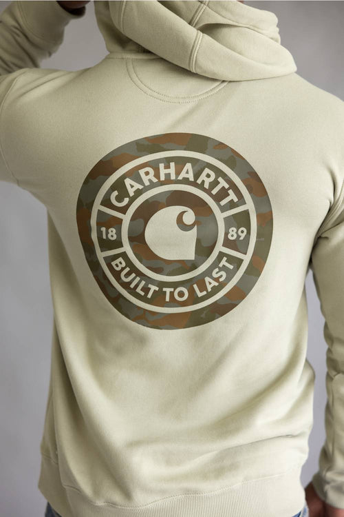 A person wearing a light beige hoodie from the back, featuring a large circular Carhartt logo printed in camouflage and white on the back.