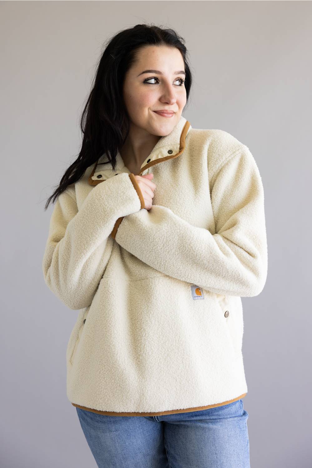 A woman wearing a cream sherpa fleece pullover jacket with tan trim and blue jeans, standing against a plain light gray background.