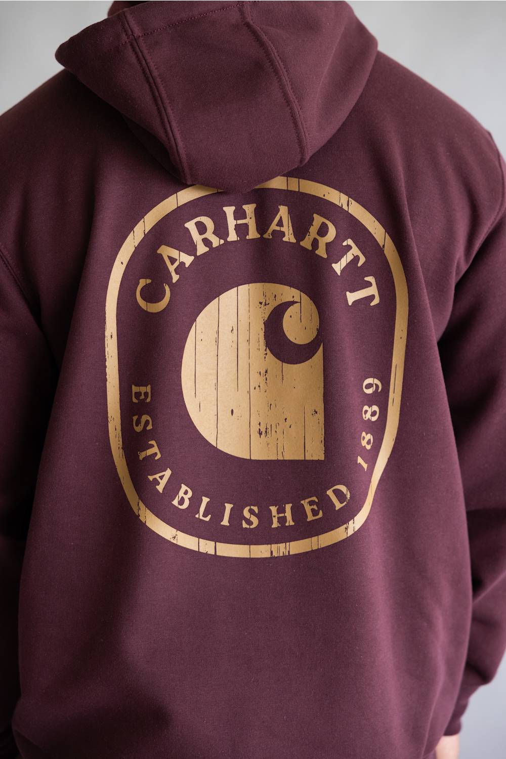A close-up of a maroon hoodie showing the back, featuring a large distressed tan graphic with the text 'Carhartt Established 1881' and the brand's signature 'C' logo.