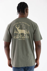 A man viewed from behind wearing a sage green Carhartt t-shirt with a large beige graphic on the back featuring a deer, trees, and the Carhartt logo.