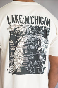 A close-up of a person wearing a cream-colored t-shirt, showing the back of the shirt with a large black graphic map of Lake Michigan and surrounding counties.