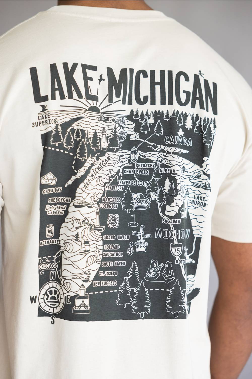 A close-up of a person wearing a cream-colored t-shirt, showing the back of the shirt with a large black graphic map of Lake Michigan and surrounding counties.