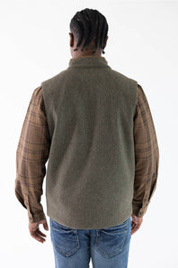 A man viewed from behind wearing an olive green vest over a brown and tan plaid flannel shirt, paired with blue jeans, against a plain white background.