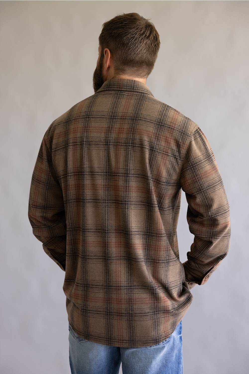 A man viewed from behind wearing a brown and beige plaid flannel shirt and blue jeans, standing against a neutral gray background.