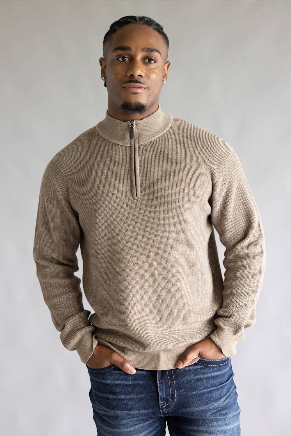 A man wearing a light beige ribbed knit quarter-zip sweater with a mock neck collar, paired with blue jeans, standing against a neutral gray studio background.
