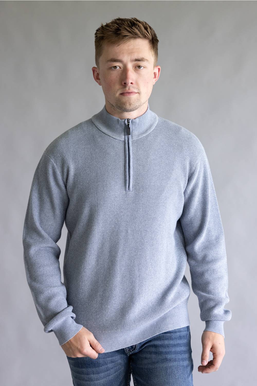 A man wearing a light gray ribbed quarter-zip sweater and blue jeans, standing against a neutral gray studio background.