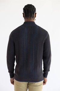 A man viewed from the back wearing a brown knit sweater with blue cable-stitch detailing down the center, paired with khaki pants, against a plain white background.