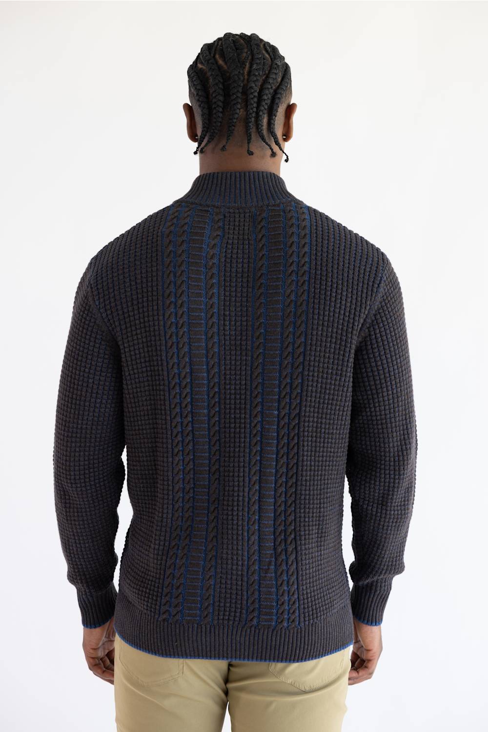 A man viewed from the back wearing a brown knit sweater with blue cable-stitch detailing down the center, paired with khaki pants, against a plain white background.