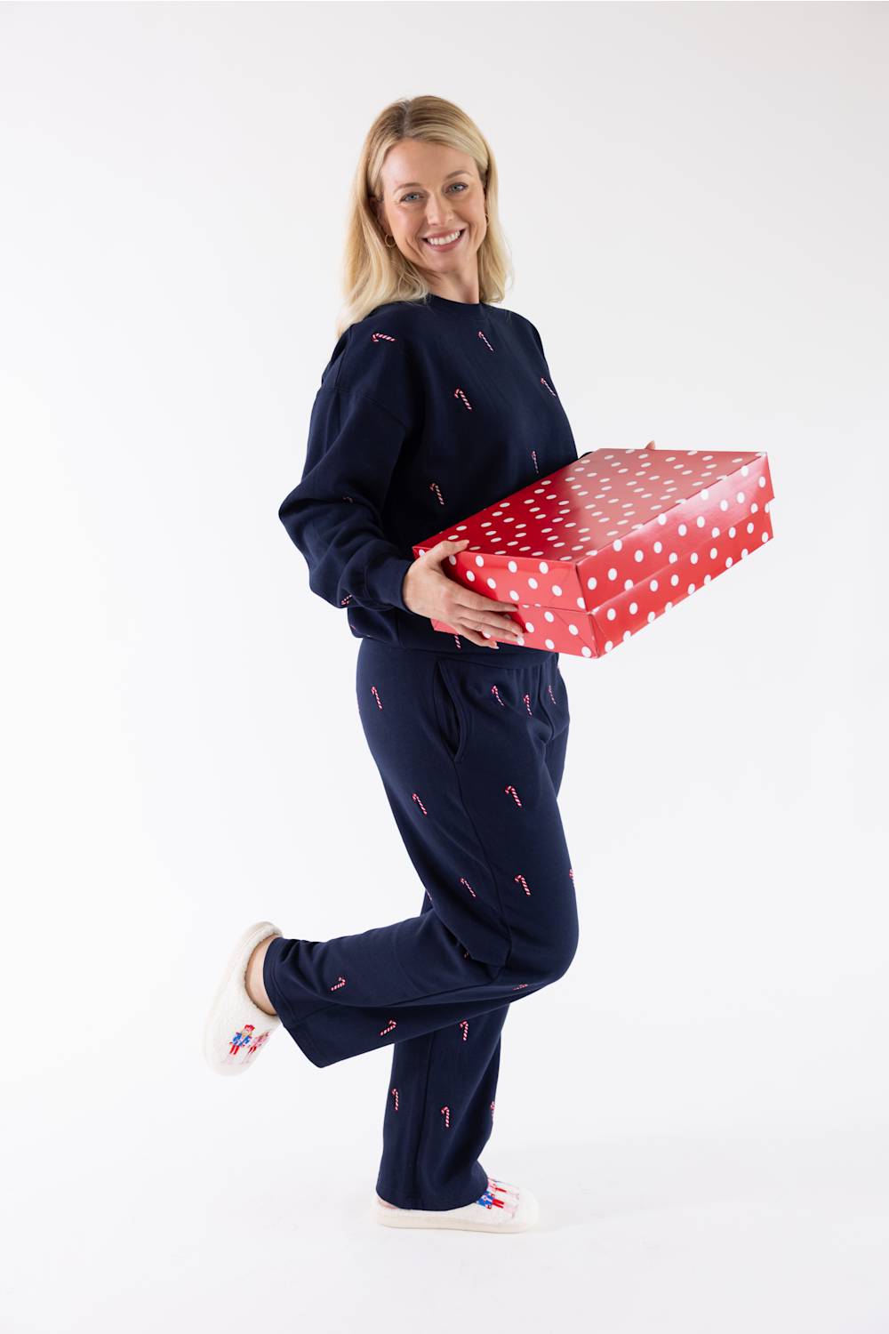 A smiling woman holding a large red and white polka-dot gift box, wearing a navy blue crewneck fleece sweatshirt and matching straight-leg sweatpants featuring an all-over embroidered red and white candy cane pattern, with white slippers