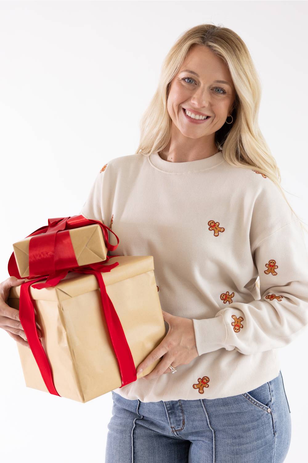 A smiling blond woman holding wrapped Christmas presents while wearing a beige fleece sweatshirt embroidered with all-over gingerbread men.