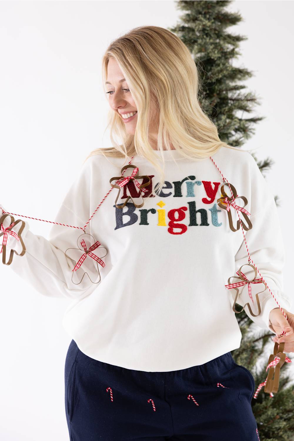 Woman wearing an ivory 1897 Active Patch Merry and Bright Fleece Sweatshirt with colorful chenille "Merry & Bright" holiday graphics, posing in front of a Christmas tree while holding a string of cookie cutters.