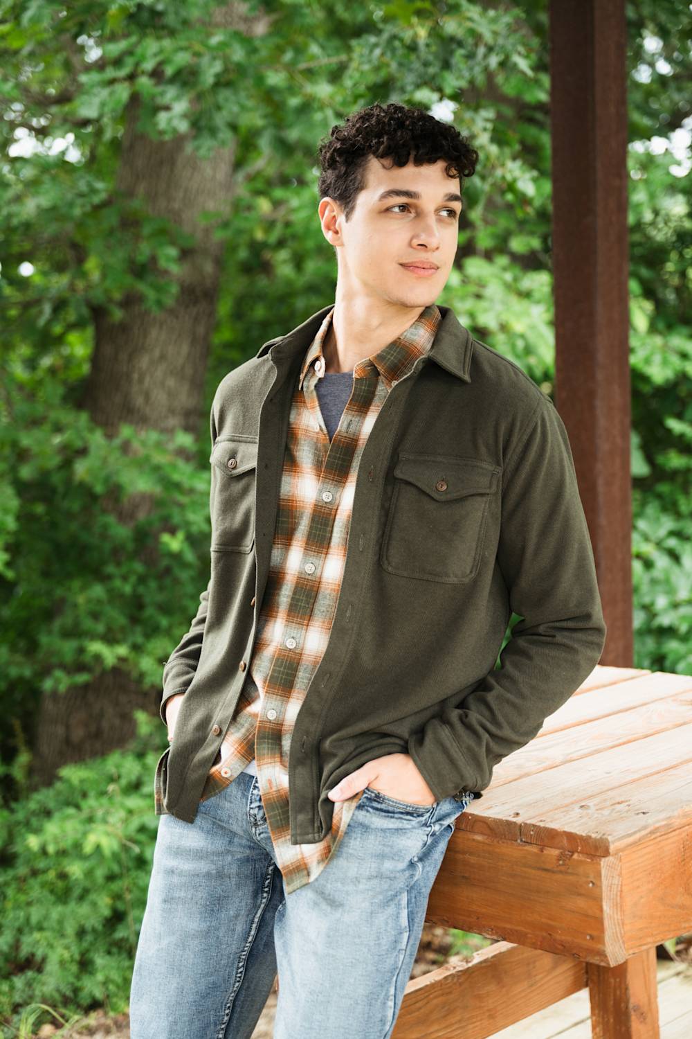 A young man wearing an olive green utility jacket over a brown and beige plaid flannel shirt, paired with blue jeans, leaning on a wooden bench in an outdoor park setting.