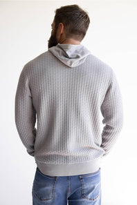 A man viewed from behind wearing a light gray waffle-knit hoodie and blue jeans against a plain white background.
