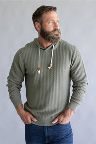 A man with a full beard wearing an olive green waffle-knit hoodie with white drawstrings, paired with blue jeans, standing against a neutral gray background.