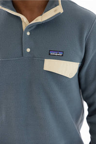 Close-up of a Patagonia fleece pullover in a muted slate blue color, featuring a cream-colored collar, placket, and chest pocket with a visible Patagonia logo patch.