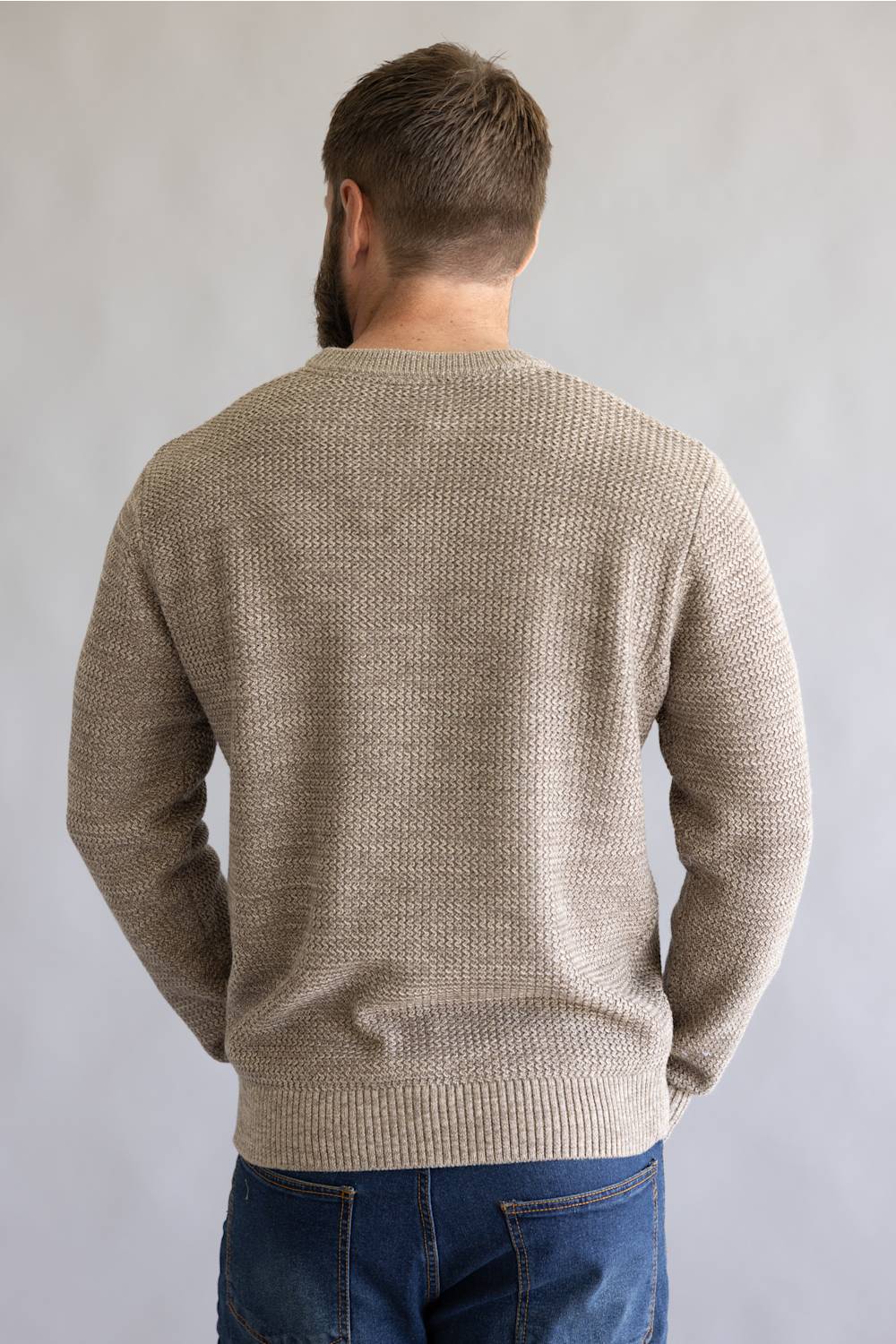 A man viewed from behind wearing a textured beige knit sweater and blue jeans against a plain light gray background.