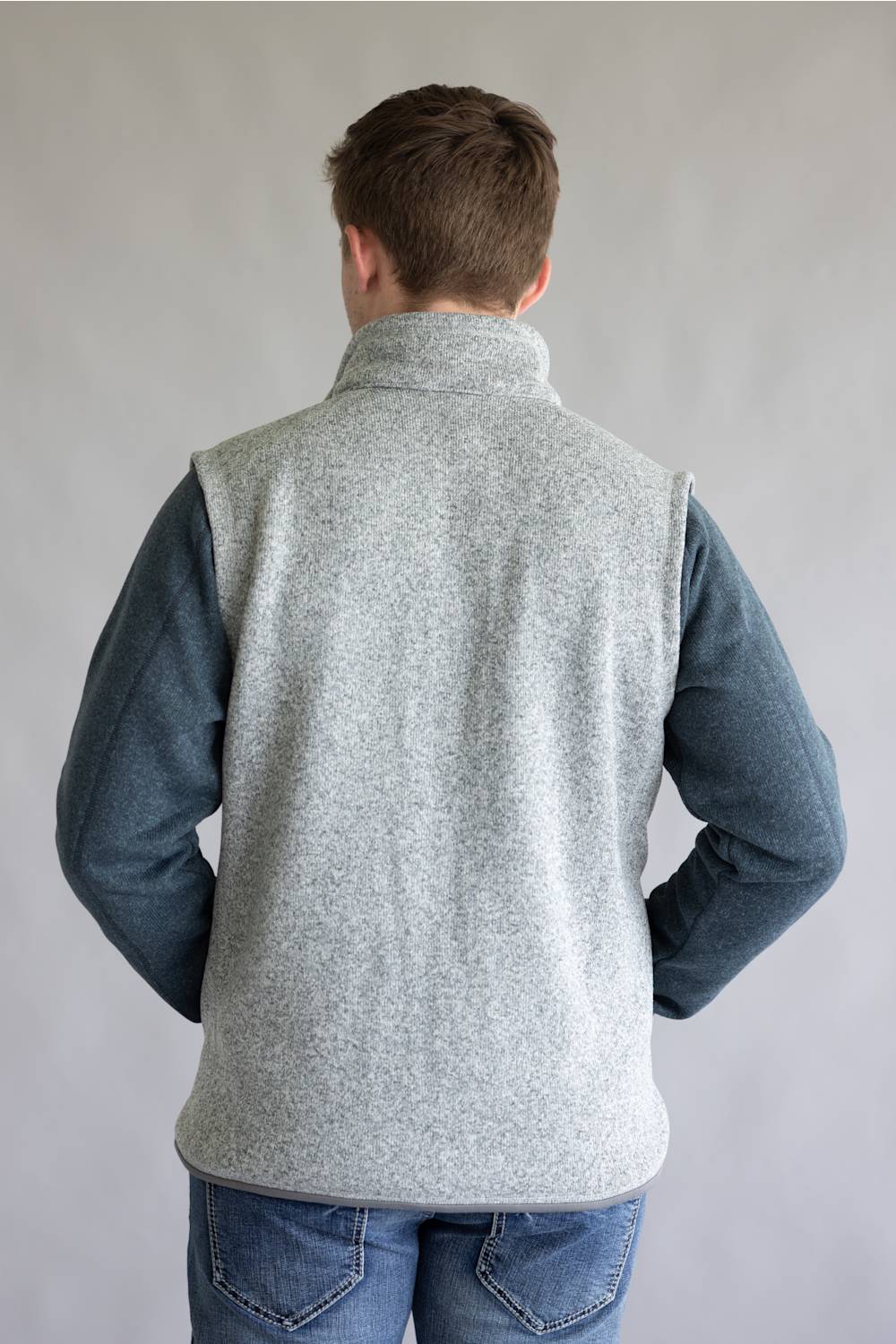 A man viewed from behind wearing a light gray heather fleece vest over a slate blue long-sleeve shirt and blue jeans, standing against a neutral gray background.
