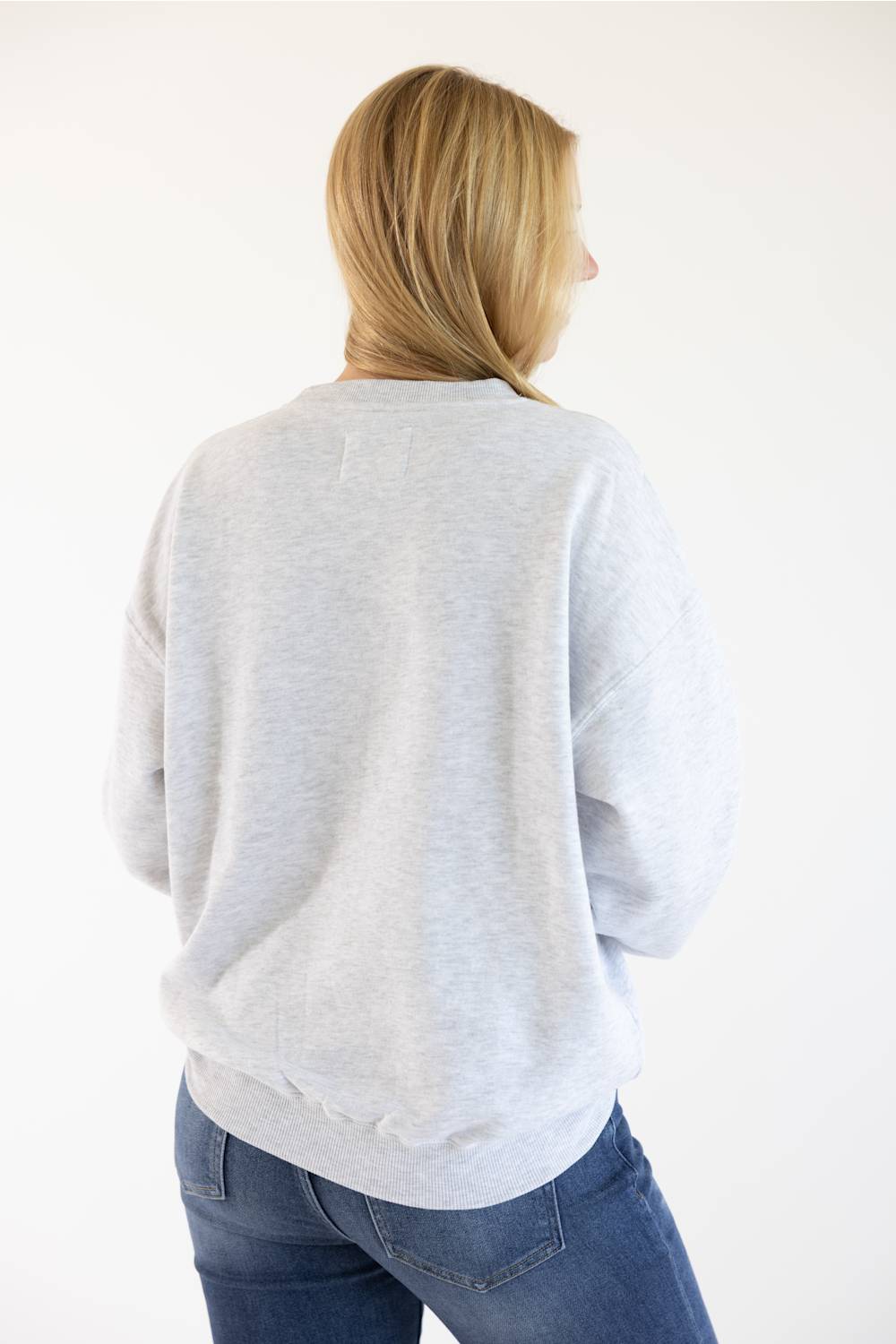 A woman with blonde hair is shown from behind wearing a light gray heather crewneck sweatshirt and blue jeans against a plain white background.