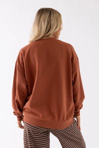 A woman viewed from behind wearing a rust-colored crewneck sweatshirt and brown-and-white striped pants against a plain white studio background.