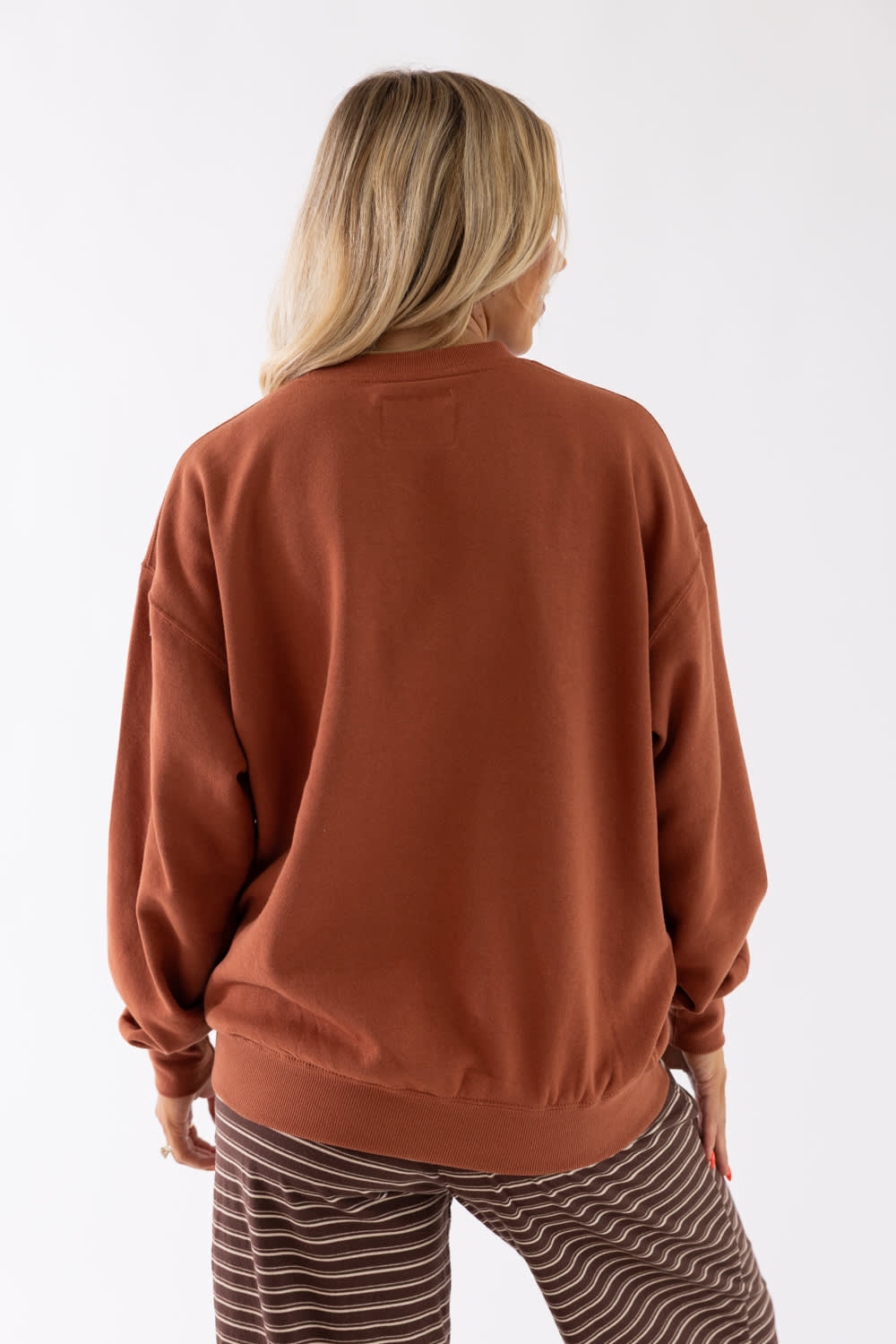 A woman viewed from behind wearing a rust-colored crewneck sweatshirt and brown-and-white striped pants against a plain white studio background.